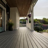 A spacious front porch features a sleek wooden deck and comfortable seating, set against a modern farmhouse exterior. Potted plants and hanging baskets add greenery, while the setting sun casts a warm glow over the tranquil outdoor space. The porch overlooks a lush garden with blooming flowers and a distant fence, creating a peaceful and inviting atmosphere.