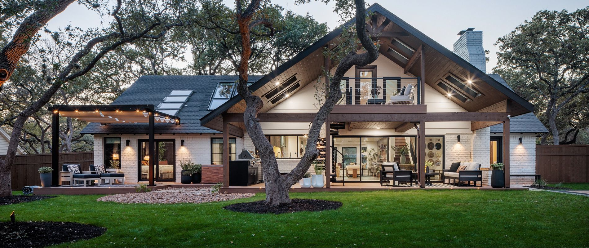 A contemporary two-story house features expansive glass windows and a spacious covered patio area. The home is set in a lush, green yard surrounded by mature trees, creating a tranquil outdoor environment. Warm exterior lighting and visible seating areas enhance the inviting atmosphere. The architectural style blends modern and rustic elements, with wood accents and clean lines. No visible text or numeric values are present in the image.
