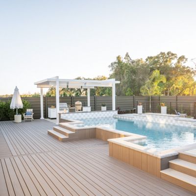 A contemporary outdoor swimming pool is the main feature of this backyard, surrounded by a large wooden deck and steps. The setting includes lounge chairs, a shaded pergola, and potted plants, all bathed in warm sunlight. The scene is tranquil and inviting, with clean lines and neutral tones enhancing the modern style. No visible text or numbers are present in the image.
