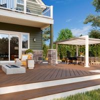 A spacious wooden patio features comfortable outdoor seating with yellow cushions and a central fire pit. The setting includes a covered pergola with a dining table, string lights, and lush greenery in the background. Large sliding glass doors lead into the house, and decorative lanterns add a cozy touch. The scene is captured on a bright, sunny day with clear blue skies. No visible text or numbers are present in the image.