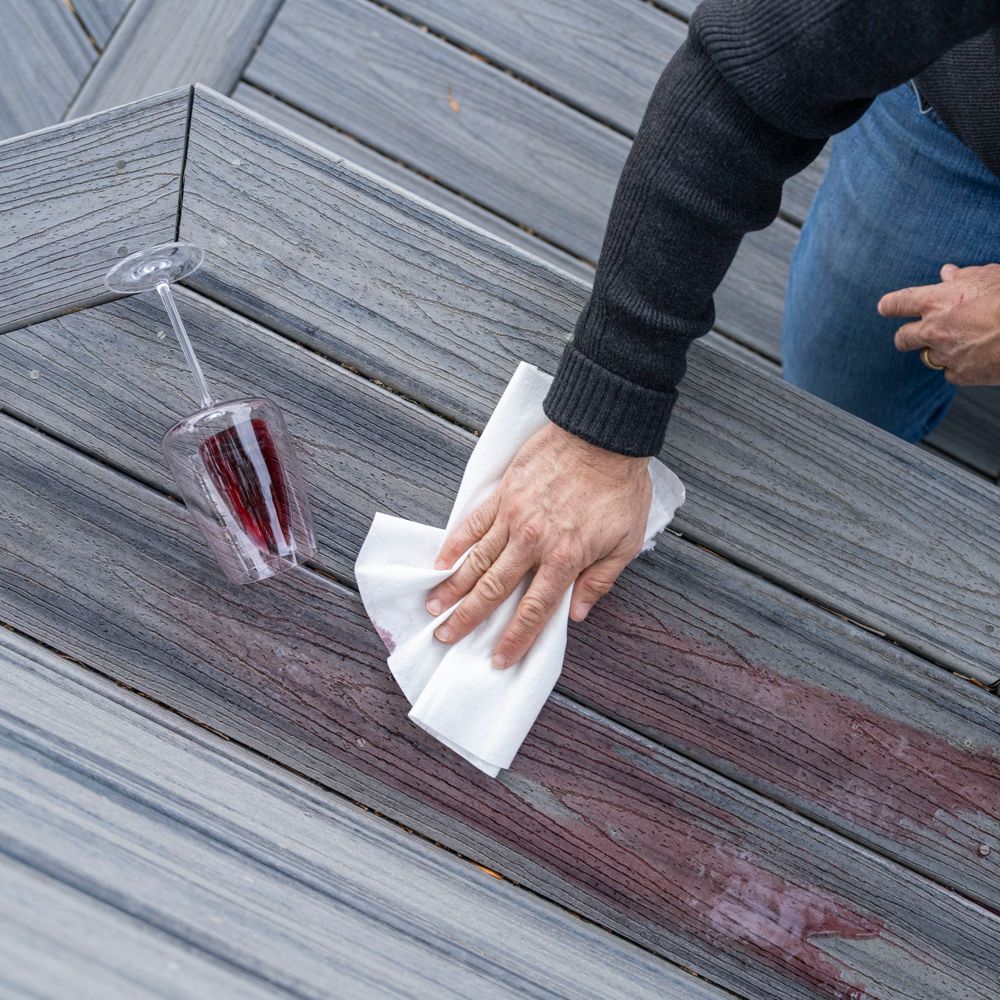 A person is wiping up a red wine spill from a gray wooden deck using a white cloth. A tipped-over wine glass lies next to the stain, indicating a recent accident. The scene is outdoors, and the person is wearing a dark sweater and jeans. The focus is on the cleaning action and the vivid contrast between the wine and the deck surface.
