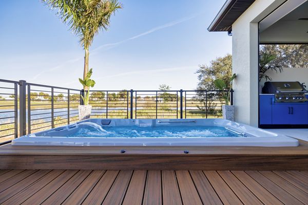 A contemporary hot tub is set into a wooden deck on a spacious outdoor patio. The scene overlooks a tranquil waterway, with palm trees and a stainless steel grill visible in the background. The setting is bright and inviting, featuring clear blue water and a clean, modern aesthetic. No people are present, emphasizing relaxation and leisure. The overall mood is serene and luxurious.