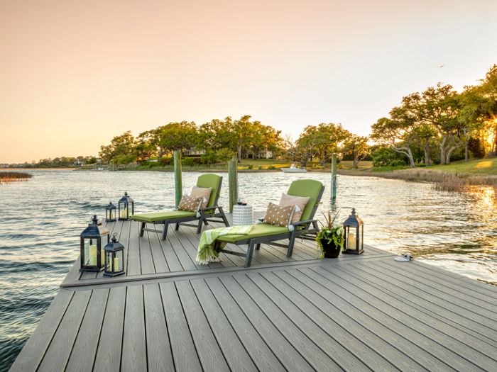 Two green-cushioned lounge chairs sit on a modern wooden dock overlooking a calm lake at sunset. Decorative lanterns and potted plants enhance the tranquil outdoor setting. The scene is peaceful, with warm golden light reflecting off the water and trees in the background.