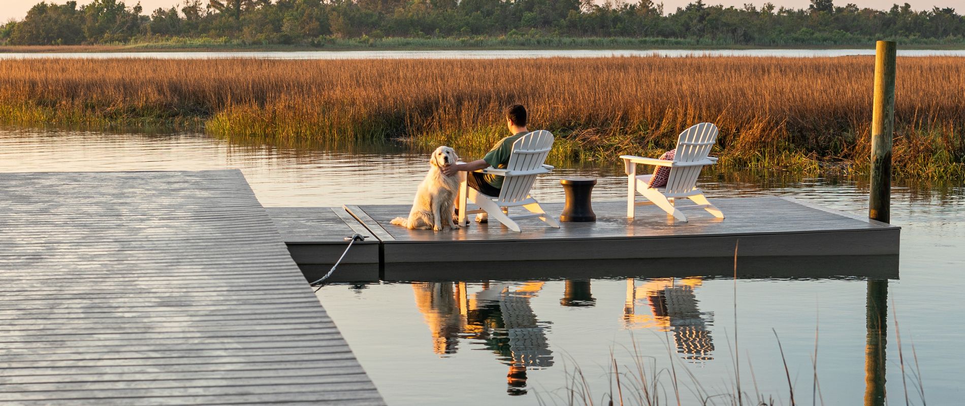 A person sits with a dog on a small dock, both facing the water during a serene sunset. Two white Adirondack chairs are positioned on the dock, surrounded by calm water and marsh grasses. The scene is tranquil, with soft golden light and reflections on the water. The setting suggests relaxation and peaceful outdoor leisure. No visible text or numbers are present in the image.