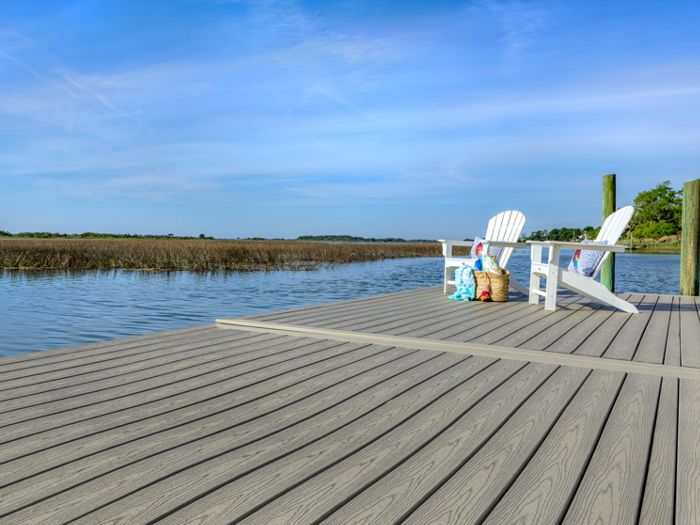 Two white Adirondack chairs sit on a spacious wooden dock overlooking a calm lake or river. The scene is set under a clear blue sky with a few decorative pillows and a tote bag adding color to the tranquil setting. Tall grasses line the water’s edge, and the dock’s textured planks are clearly visible. The overall mood is serene and inviting, perfect for relaxation.