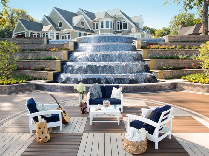 A stylish outdoor seating area with white and navy cushioned chairs surrounds a coffee table on a circular wooden deck. The setting overlooks a dramatic multi-level waterfall with a large, elegant house in the background. Decorative items like a guitar, vase of flowers, and cozy blankets add a relaxed, inviting atmosphere. The scene is bright and tranquil, with lush landscaping and clear skies. No visible text or numbers are present in the image.