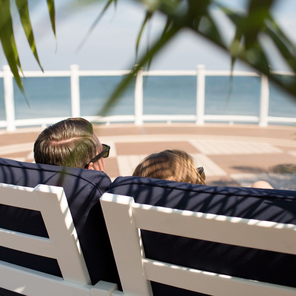 A man and woman sit closely together on white outdoor chairs, facing the ocean. The setting is a sunny seaside deck with a clear view of the water, framed by a white railing and tropical leaves in the foreground. The mood is relaxed and tranquil, with soft natural lighting and a calm atmosphere. Both individuals appear to be adults, enjoying a peaceful moment by the sea.