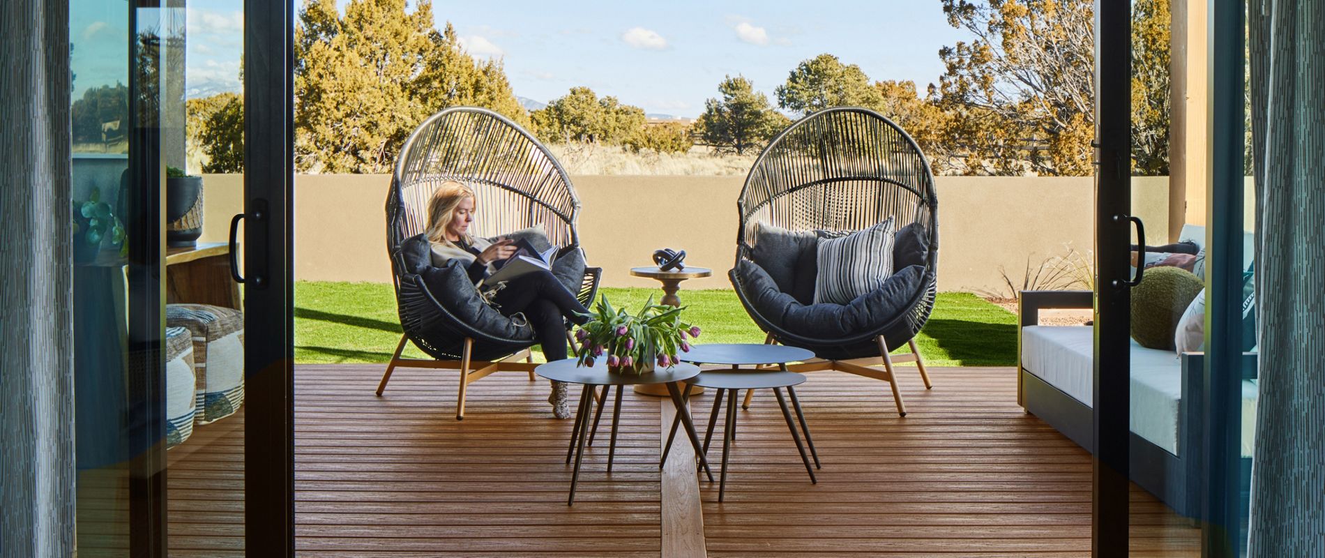 A woman sits comfortably in a round, cushioned chair on a wooden patio deck, viewed through open glass doors. The outdoor setting features contemporary furniture, a small table with plants, and a backdrop of trees and a privacy wall. The scene is bright and tranquil, with natural light and a relaxed atmosphere.