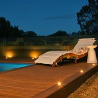 A contemporary wooden pool deck features a cushioned lounge chair and a small side table, illuminated by warm recessed lights. The setting is outdoors at dusk, with a tranquil blue sky and soft landscape lighting enhancing the relaxing atmosphere. The pool edge and surrounding greenery add to the serene, upscale ambiance. No visible text or numbers are present in the image.