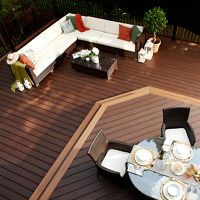 A spacious wooden deck features a cozy sectional sofa with colorful pillows and a coffee table, set in a lush outdoor environment. In the foreground, a dining table is elegantly set for a meal with plates, glasses, and decorative accents. The scene is bright and inviting, with natural wood tones and greenery enhancing the relaxed, contemporary atmosphere. No visible text or numbers are present in the image.