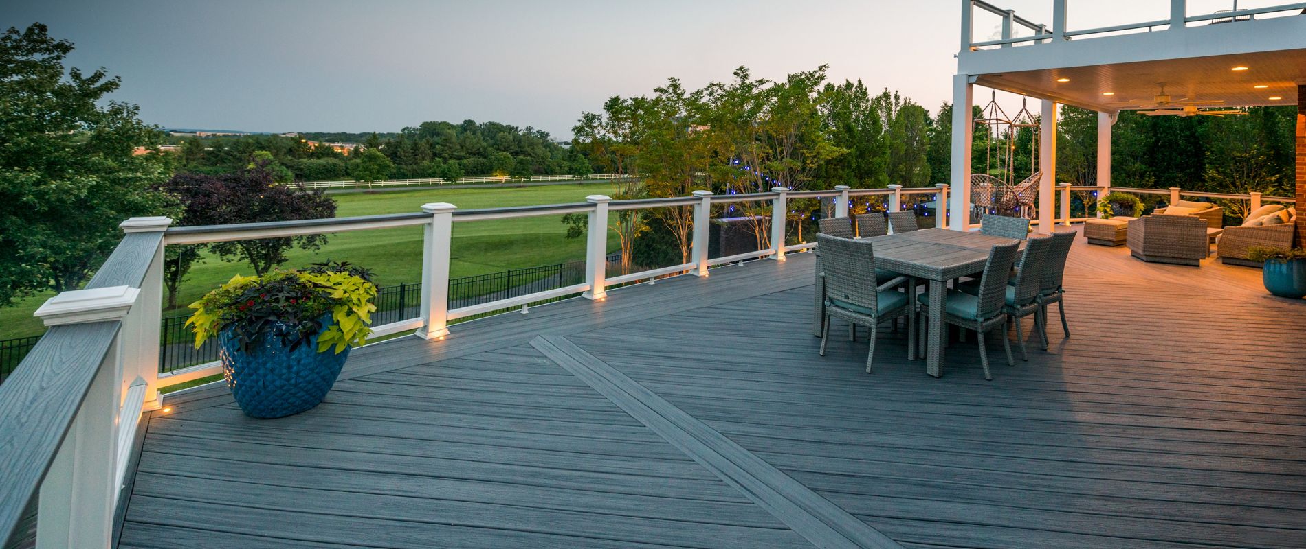 A large, contemporary outdoor deck features sleek gray flooring and glass railings, offering an expansive view of a lush green field. The setting includes a dining table with chairs and a covered lounge area with comfortable seating. Soft ambient lighting lines the deck, and large potted plants add a touch of nature. The tranquil evening sky enhances the relaxed, inviting mood.
