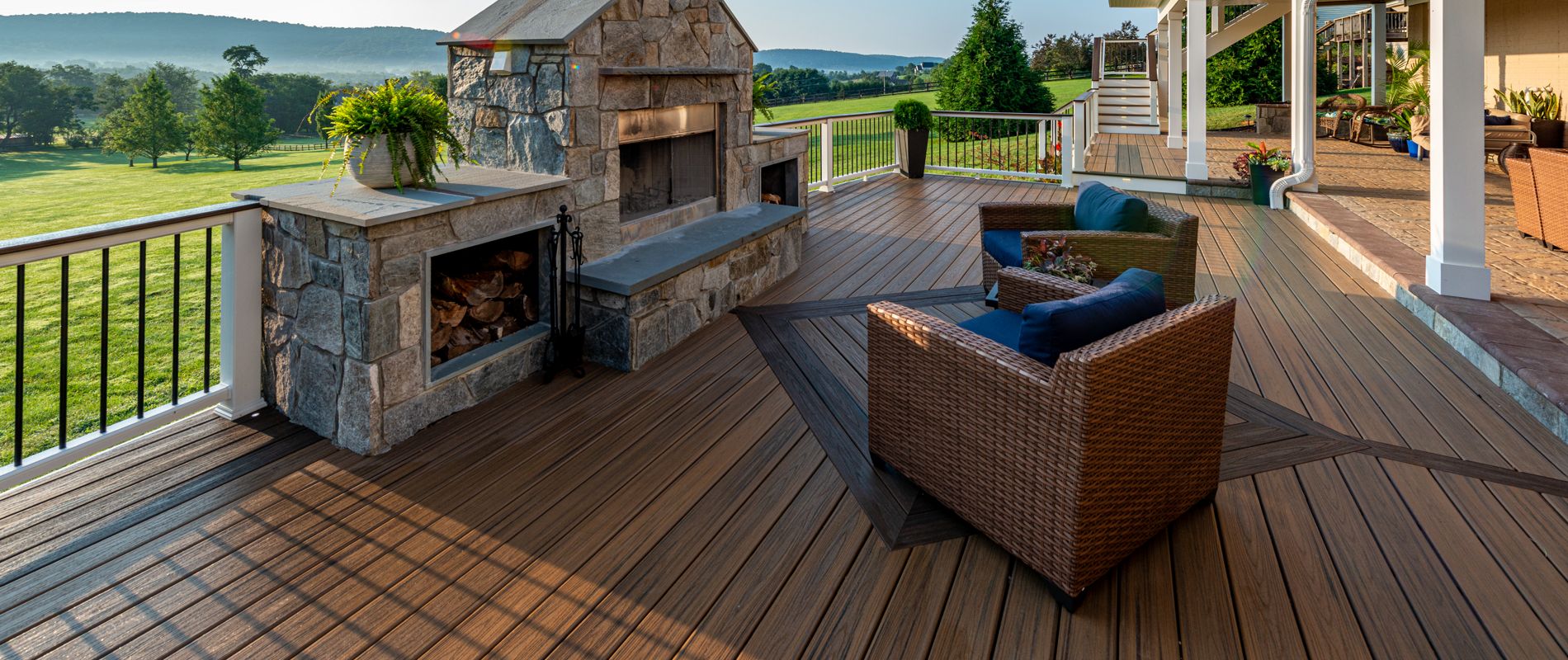 A large outdoor deck features a prominent stone fireplace and comfortable wicker seating. The setting overlooks a lush green field and rolling hills under a clear blue sky, creating a serene and inviting atmosphere. The deck is accented with potted plants and modern railings, with warm wood tones throughout. No visible text or numbers are present in the image.