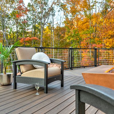 A contemporary outdoor patio features cushioned seating and a rectangular fire pit as its centerpiece. The setting overlooks a vibrant forest with colorful autumn leaves, creating a cozy and inviting atmosphere. A glass of white wine sits on the deck, and a potted plant adds a touch of greenery. The scene is bathed in warm, natural light, emphasizing relaxation and comfort.