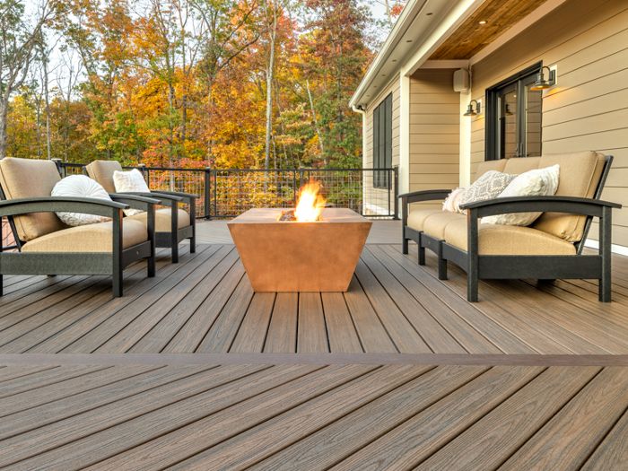 A spacious outdoor deck features a central fire pit surrounded by cushioned armchairs. The setting is a residential backyard with autumn trees in the background, creating a cozy and inviting atmosphere. The deck boards are arranged in a chevron pattern, and the neutral color palette enhances the modern style. No visible text or numbers are present in the image.