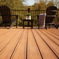 A close-up view of a wooden deck highlights the natural grain and warm tones of the planks. In the background, two Adirondack chairs and a small table create a cozy seating area, with a blanket draped over one chair. The scene is set outdoors, surrounded by trees and soft sunlight, evoking a peaceful and inviting atmosphere.