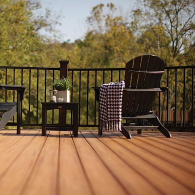 A spacious wooden deck features two black Adirondack chairs facing a scenic view of trees. A small black side table sits between the chairs, holding a mug, a small plant, and a book. A checkered blanket is draped over one chair, adding a cozy touch to the tranquil outdoor setting. The scene is bathed in warm, natural light, highlighting the earthy tones of the deck.