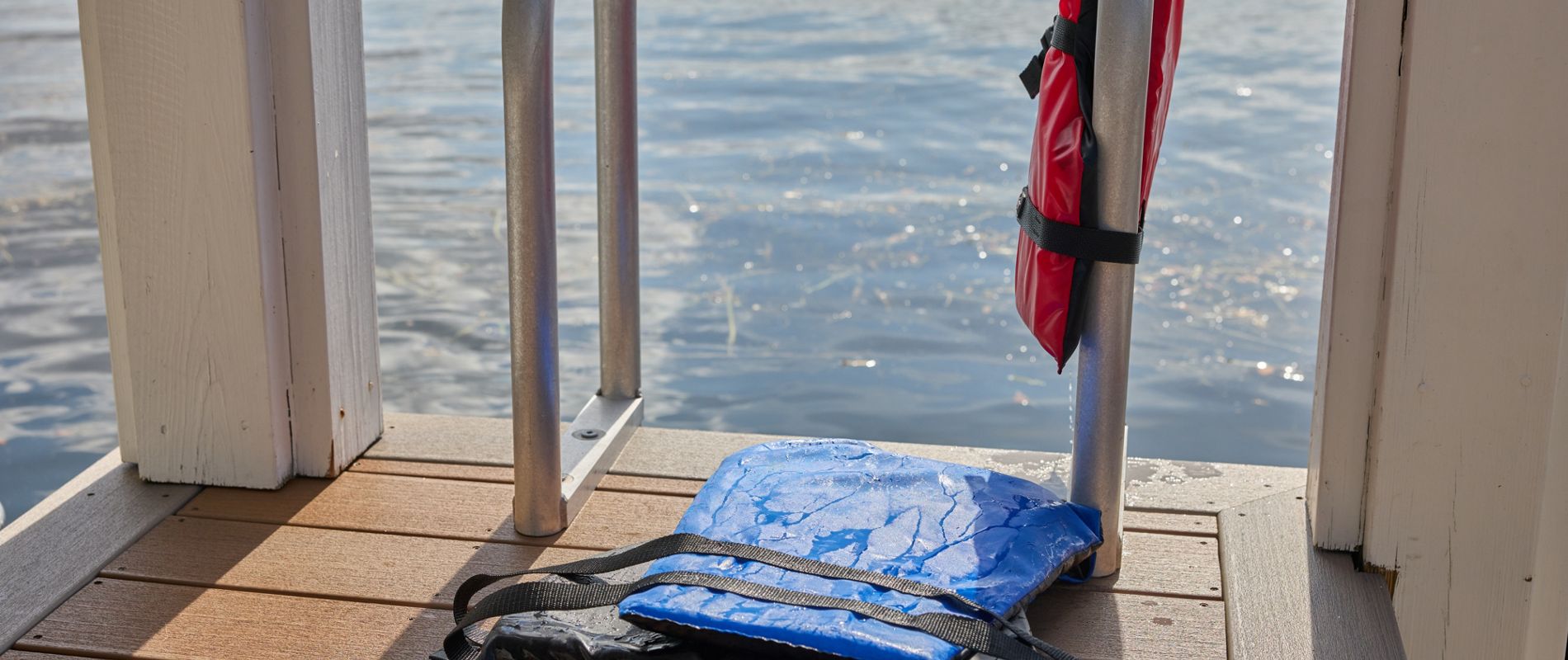 A blue life jacket lies on a wooden dock next to a metal ladder, with a red life jacket hanging on the railing. The scene overlooks calm lake water, suggesting a peaceful outdoor setting. The sunlight highlights the wet surface of the blue jacket, indicating recent use. No visible text or numbers are present in the image.