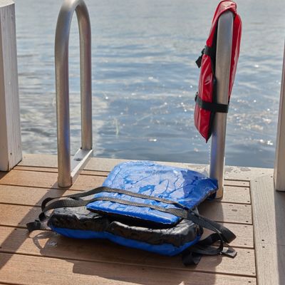 A blue life jacket lies on a wooden dock next to a metal ladder, with a red life jacket hanging on the railing. The scene overlooks calm lake water, suggesting a peaceful outdoor setting. The sunlight highlights the wet surface of the blue jacket, indicating recent use. No visible text or numbers are present in the image.