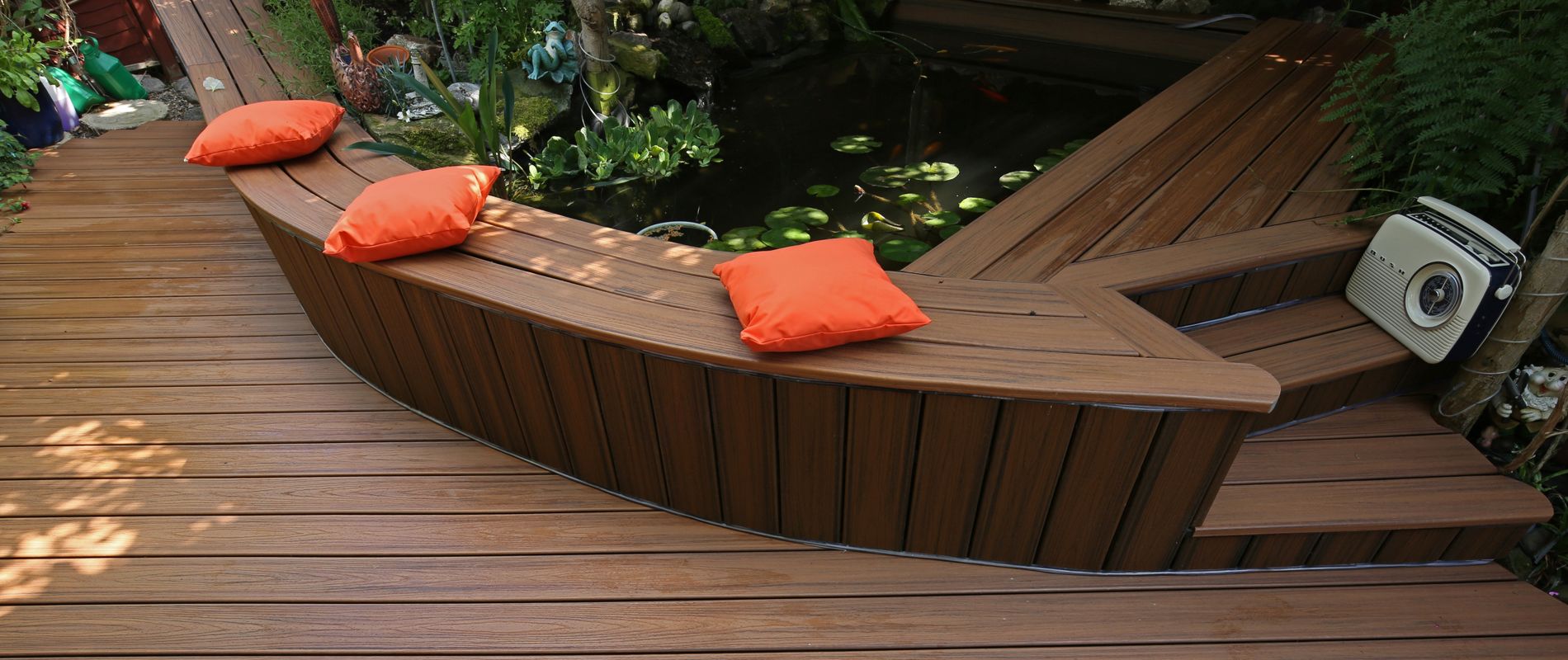 A contemporary wooden deck features clean lines and a built-in seating area with three bright orange cushions. The deck overlooks a tranquil garden pond filled with lily pads and surrounded by lush greenery. A small portable speaker sits on the steps, adding a touch of leisure to the outdoor setting. The overall mood is relaxed and inviting, with natural wood tones and pops of color.