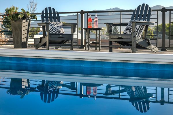 Two black Adirondack chairs with patterned cushions are arranged beside a swimming pool, separated by a small table holding two bottles and glasses. The setting is outdoors on a sunny day, with mountains visible in the background and a sleek metal railing enclosing the patio. The scene reflects a relaxed, contemporary mood with a blue and neutral color palette, and the pool water mirrors the furniture above.