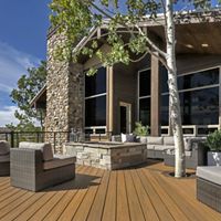 A spacious wooden deck features contemporary wicker lounge chairs and a central stone fire pit. The setting is an outdoor patio attached to a modern house with large windows and stone accents. Trees and scenic hills are visible in the background, creating a relaxed and inviting atmosphere. The color palette includes natural wood tones, gray cushions, and green foliage.