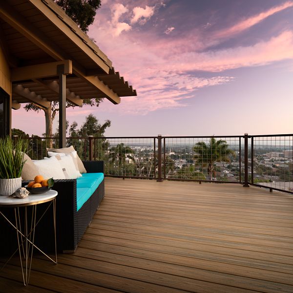 A spacious wooden deck patio features a contemporary outdoor sofa with turquoise cushions and a side table with fruit. The setting overlooks a cityscape with lush trees and palm trees, under a vibrant sunset sky with pink and purple hues. Decorative plants add a touch of greenery, creating a tranquil and inviting atmosphere.