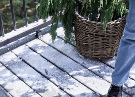 A person in jeans and dark shoes walks across a wooden deck lightly dusted with snow. A large wicker basket with evergreen branches sits nearby, adding a touch of greenery to the wintry scene. The setting appears to be an outdoor patio or balcony, with a black metal railing visible. The overall mood is calm and seasonal, with natural textures and cool tones.