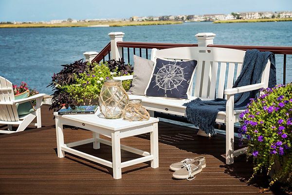 A white wooden bench with nautical-themed pillows and a blue throw sits on a deck overlooking a river or bay. A matching white table holds glass lanterns and a book, while potted flowers and plants add color to the scene. The setting is bright and inviting, with a relaxed, coastal mood and visible residential buildings in the distance.