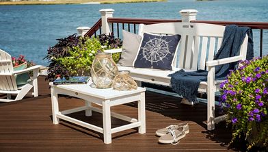 A white wooden bench with nautical-themed pillows and a blue throw sits on a deck overlooking a river or bay. A matching white table holds glass lanterns and a book, while potted flowers and plants add color to the scene. The setting is bright and inviting, with a relaxed, coastal mood and visible residential buildings in the distance.