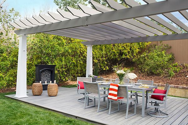 A contemporary backyard patio features a sleek gray pergola over a wooden deck. The setting includes a dining table with chairs, accented by red and orange cushions and a striped towel. Decorative elements like woven stools and fresh flowers add a welcoming touch, while lush greenery surrounds the space. The area is designed for outdoor dining and relaxation.