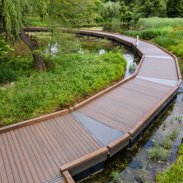 A gently curving wooden boardwalk stretches through a vibrant green wetland area, bordered by tall grasses and trees. The path is elevated above shallow water, creating a tranquil and inviting scene. A single person stands in the distance, adding a sense of scale and peaceful solitude to the natural setting.