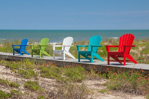 A row of five Adirondack chairs in vibrant colors sits on a wooden boardwalk overlooking a sandy beach and calm ocean. The chairs are blue, green, white, turquoise, and red, creating a cheerful and inviting scene. Tall grasses and wildflowers grow around the boardwalk, with the sea and blue sky in the background. The setting suggests a relaxed, summery mood perfect for leisure and vacation.