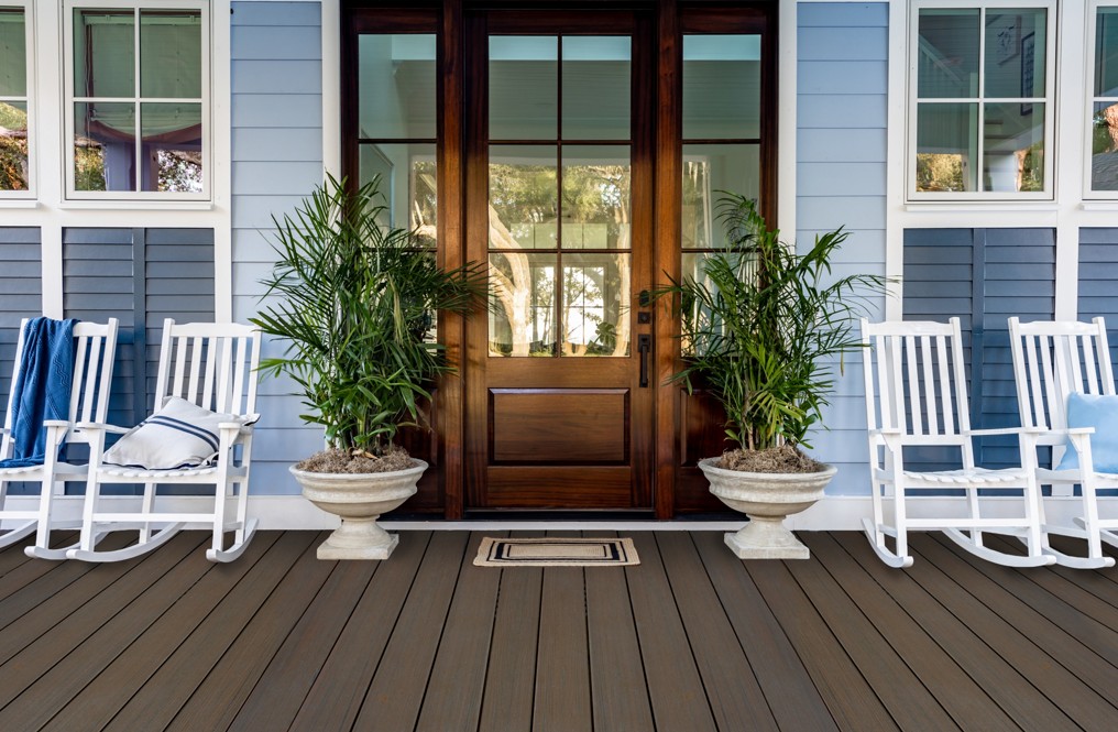 A welcoming porch featuring white rocking chairs and lush green potted plants. The setting includes a wooden door framed by glass panels and a light blue exterior wall. The wooden deck adds warmth to the scene, complemented by soft cushions and a folded blanket. The space evokes a serene and inviting atmosphere.
